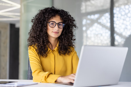 A beautiful woman with curly hair and glasses is working on a laptop in a modern office setting.の写真素材