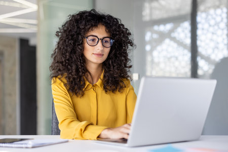 A woman with curly hair, wearing glasses and a yellow shirt, works on a laptop in an office setting. She appears focused and engaged in her tasks at the white desk.の写真素材