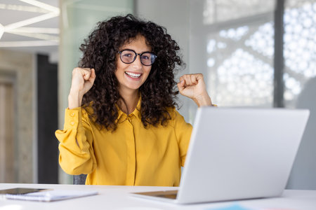 A smiling woman with curly hair wearing glasses celebrates a success while using a laptop computer.の写真素材