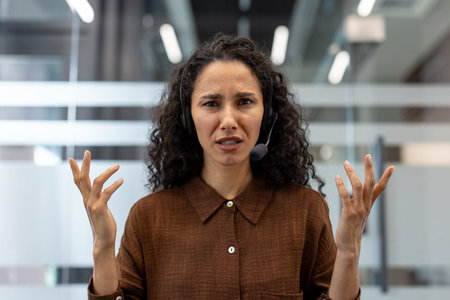 A concerned customer service representative wearing a headset is gesturing with her hands,...