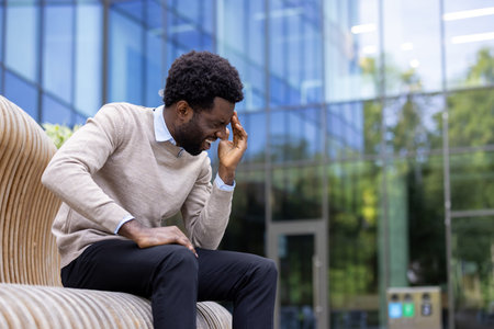 A man sits outdoors, expressing a headache, stress or pain in front of an office building, grasping...