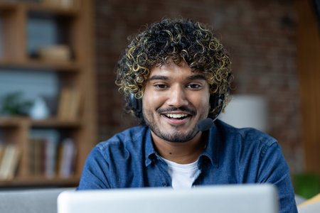 Smiling man with curly hair wearing a headset, working from home. He is looking at the camera with a...