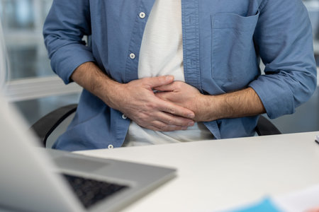 A man is seated at a desk, holding his stomach, possibly indicating abdominal pain or discomfort.の写真素材