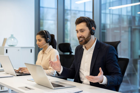 A man in a suit enthusiastically gestures during a video call, while a woman works at a computer in...