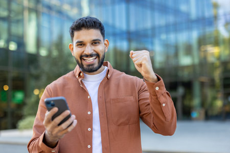 An excited man celebrating with his fist raised while holding a smartphone outside a modern office building.の写真素材