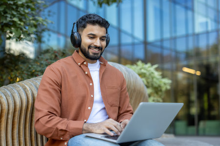 An Indian man wearing headphones smiles while using a laptop outdoors on a bench, surrounded by...