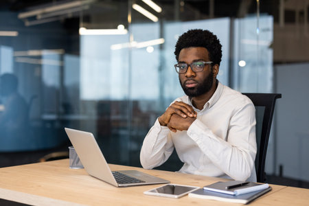Man wearing glasses in a professional setting, working with a laptop on a desk.