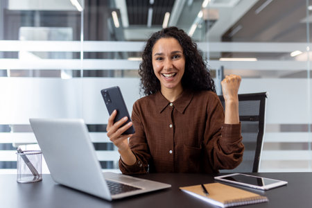 A businesswoman feeling accomplished while using her smartphone and laptop in a modern office.の写真素材