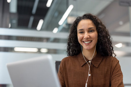 A cheerful professional woman engaging in remote work, using a laptop and earphones in an office...