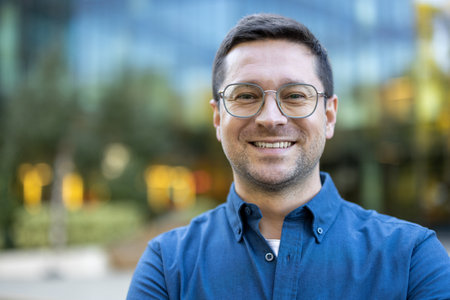 A cheerful man with glasses wearing a blue shirt stands outdoors, smiling confidently. The blurred urban setting behind him conveys both modernism and an approachable demeanor.の写真素材