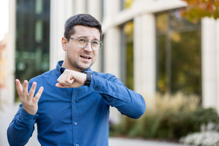A man wearing glasses and a blue shirt interacts with a smartwatch, gesturing animatedly while...