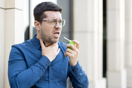 A man in a blue shirt uses a throat spray for relief while standing outside in a city setting. The...