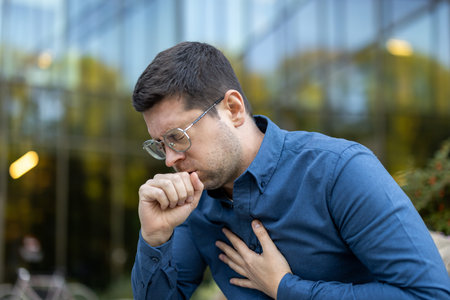 A man, appearing unwell, holds his chest and coughs, standing in front of a modern building with...