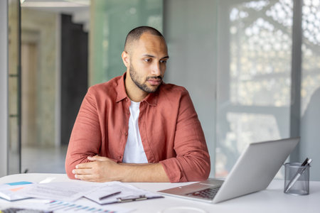 Focused young man wearing a casual shirt working remotely on his laptop in a modern environment with...
