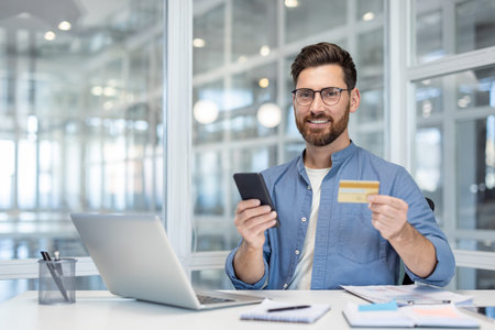 Business professional holding a smartphone and a credit card, working at a bright office desk.