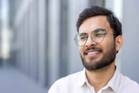 A young man confidently smiles while wearing glasses, exuding happiness and relaxation in an urban...