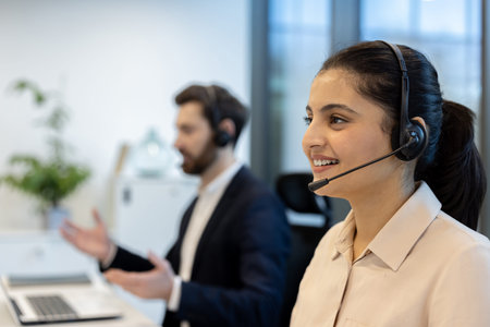 A smiling woman wearing a headset works at a call center alongside a colleague, both engaged in a...