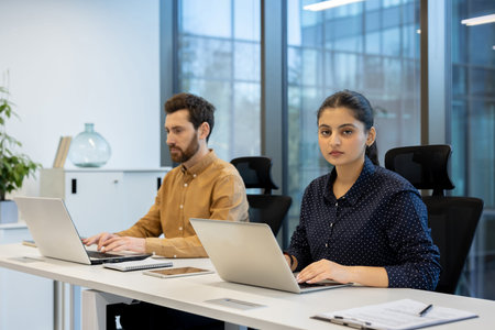 Two colleagues work at a desk using laptops in a modern office setting.