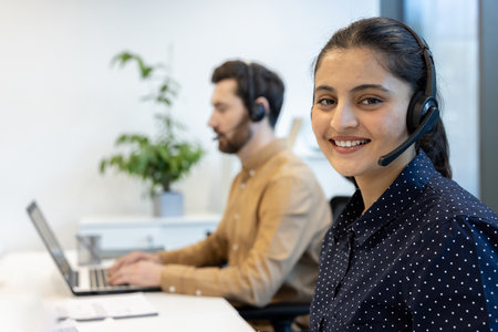 A woman with a headset smiles at the camera while working in a customer service environment, with a colleague in the background.の写真素材