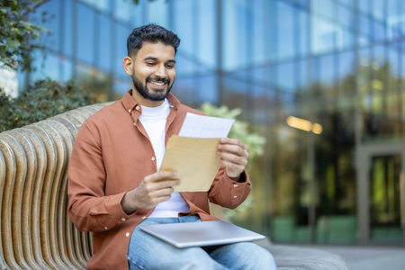 A happy man smiles reading letter outdoors, sitting on modern bench. The background includes a...
