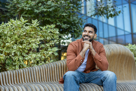 Relaxed man sitting on a stylish bench outdoors, smiling amid vibrant plants and buildings.の写真素材