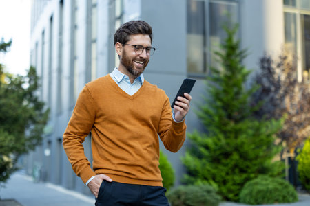 A happy man in an orange sweater looks at his smartphone while walking in a city.
