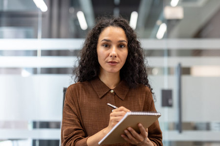 Focused business professional in a modern office environment writing notes on a notepad. Represents...