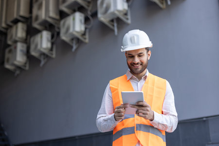 A smiling construction worker in a hard hat and reflective vest works with a tablet. The person...