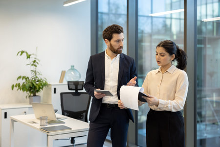 Two colleagues discuss work documents in a bright, modern office environment emphasizing...
