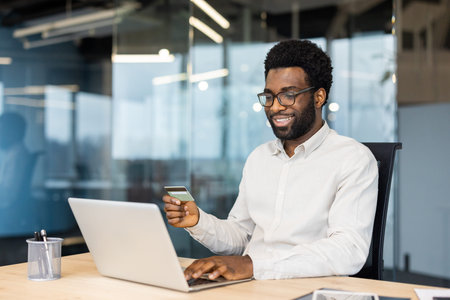A man is using his laptop and credit card for online shopping while sitting in a modern office environment. The scene highlights technology, financial transactions, and workspace aesthetics.の写真素材