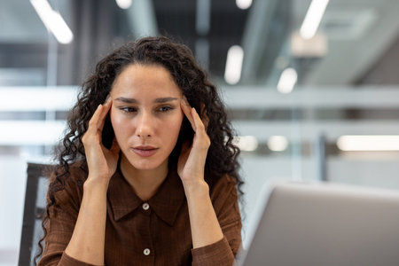 A businesswoman sits at a desk, appearing stressed and engaged in a task. The image conveys emotions...