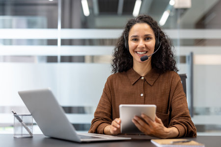 Friendly customer service agent wearing a headset while working with a tablet and laptop in a contemporary office setting, showcasing professionalism and effective communication in a workplace.の写真素材