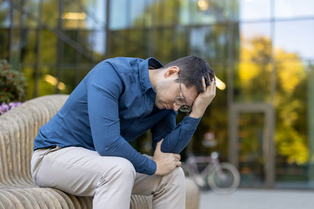 A man in despair sits on an outdoor bench, head bent, showing frustration. The glass building background highlights his urban environment, manifesting feelings of stress or concern.の写真素材
