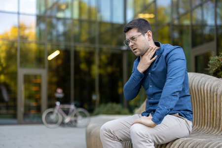 A man is seated outdoors in front of a contemporary building, clutching his throat, appearing distressed. The setting suggests an urban environment with an emphasis on health or medical urgency.の写真素材