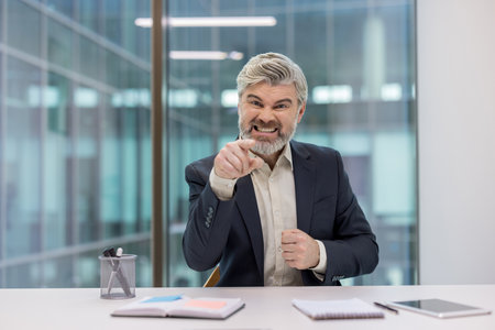 Businessman in formal attire expressing anger and frustration while pointing in an office setting.の写真素材