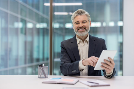 Confident middle-aged professional using a tablet at a modern office desk with joy. Portrays thoughtfulness, technology use, and the workplace environment, conveying professionalismの写真素材