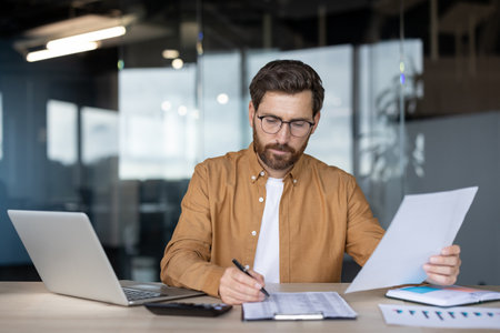 A businessman at his desk reviewing documents while working on a laptop, surrounded by charts, papers, and a calculator.の写真素材