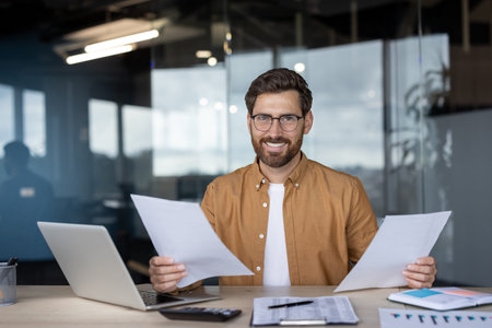 Happy businessman smiling while reviewing paperwork and working on a laptop at his desk in a modern...