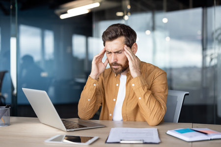 Businessman having headache and touching temples while sitting at workplace with laptop and...