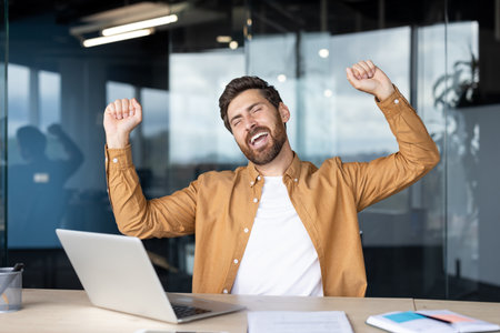 A happy man celebrates a win at his office desk, laptop open, hands raised in excitement and joy.の写真素材