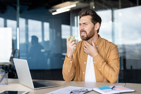 A man in an office uses a throat spray, potentially for a sore throat or cold, near a laptop.の写真素材