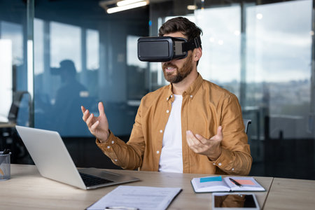 Office worker using vr headset is interacting with virtual reality in modern office, gesturing with his hands while sitting at desk with laptop and documentsの写真素材
