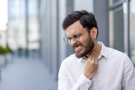Young professional man experiencing discomfort and pain while touching his neck, indicating a sore...