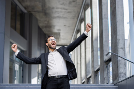 Young businessman is raising his arms in celebration, expressing joy and excitement over a recent achievement in front of an office buildingの写真素材