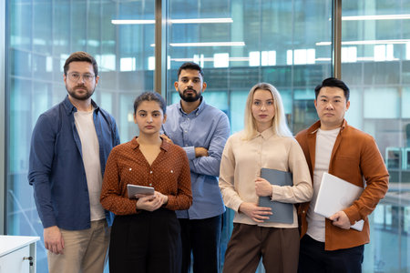 Five serious multi-ethnic businesspeople are standing in a modern office holding digital tablet,...