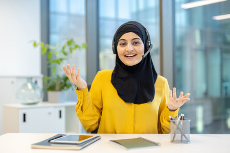 Friendly female customer service representative wearing headset and hijab explaining something using gestures while working at her desk in modern officeの写真素材