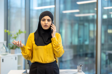 Young businesswoman wearing hijab having a difficult phone conversation in a bright modern office, gesturing with one hand while holding the phone with the otherの写真素材