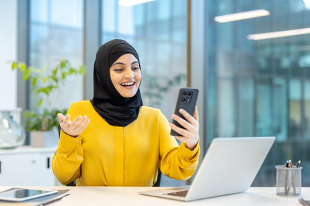 Young cheerful muslim businesswoman in hijab having video call on smartphone while sitting at desk in modern office, shes gesturing and smilingの写真素材