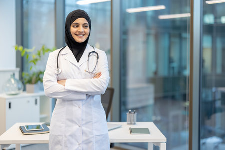 Muslim female doctor in a hijab and lab coat, confidently standing with arms crossed in a modern office, smiling and exuding professionalism in healthcareの写真素材