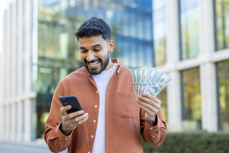 Excited businessman holding dollar bills and using smartphone app outside office building,...